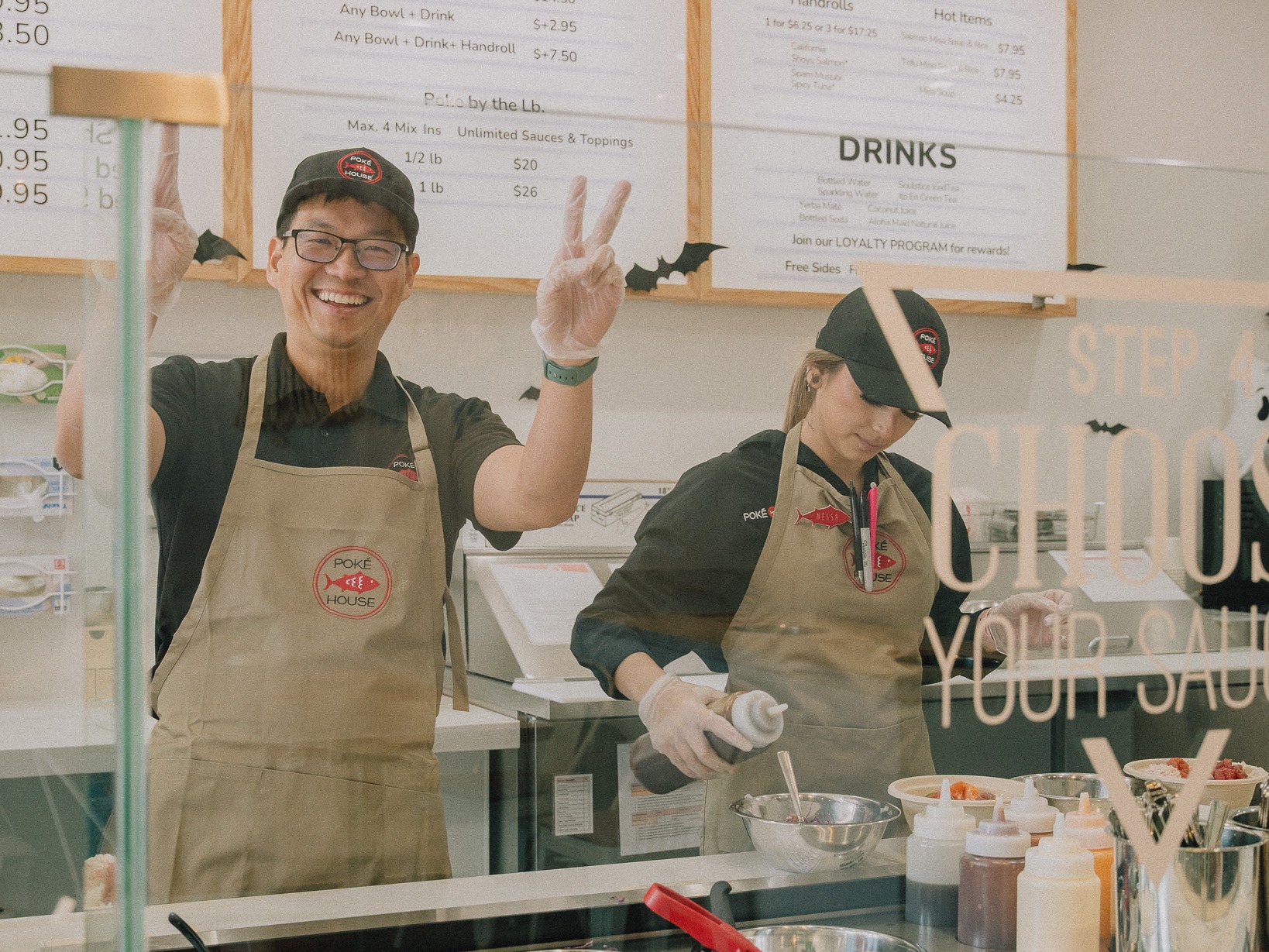 Two smiling workers behind a poke bowl counter, one making a peace sign, with colorful ingredients in the foreground and a menu board in the background.