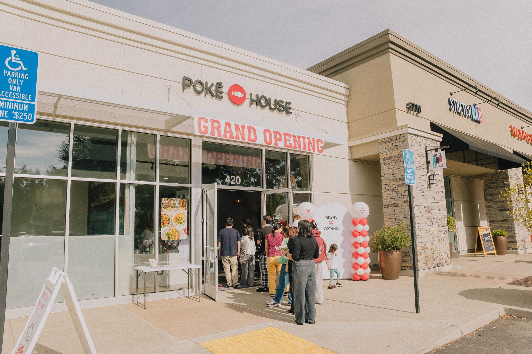 People stand in line outside a building with a "Grand Opening" sign, under the "Poke House" restaurant logo.