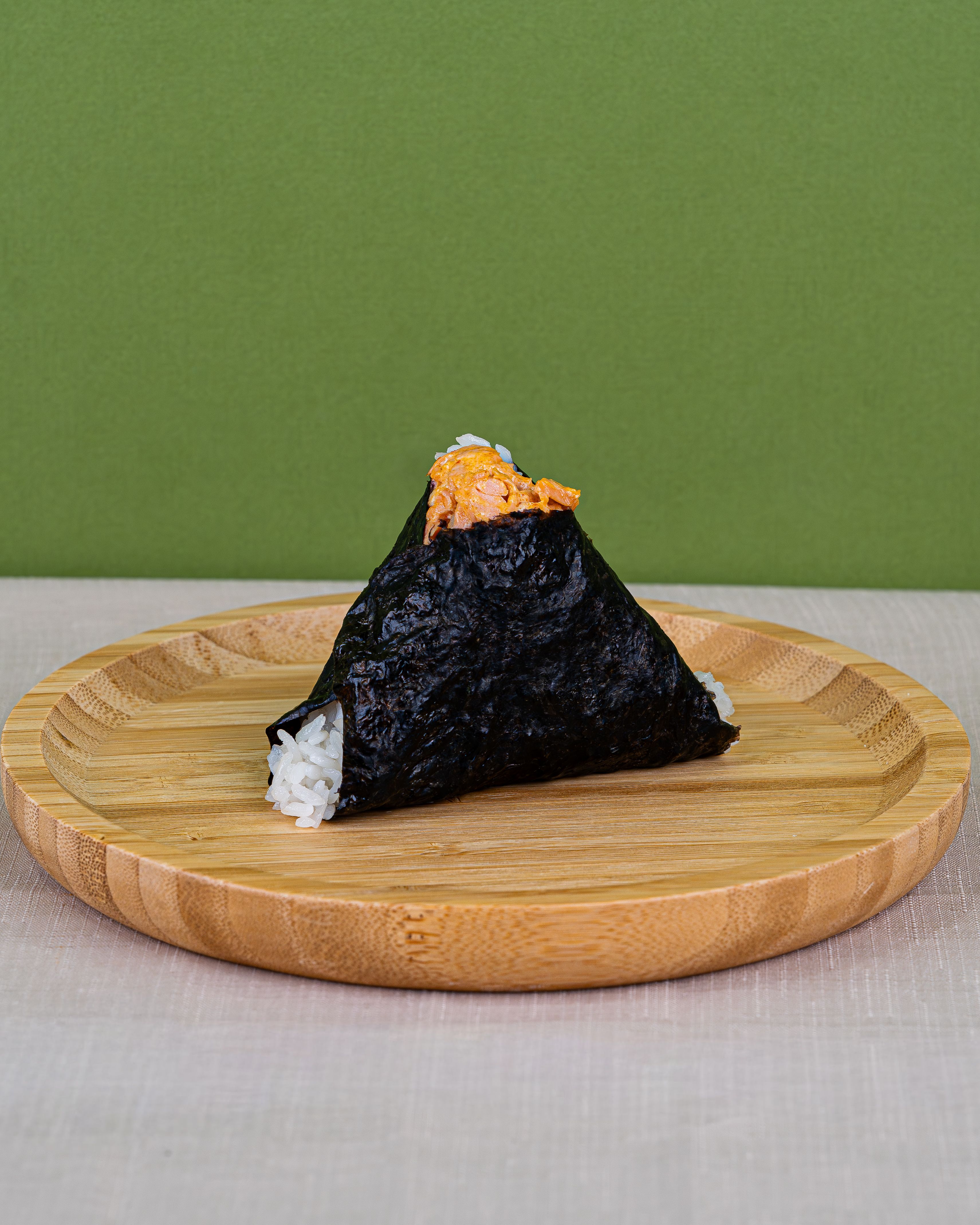 A triangular onigiri wrapped in nori with a spicy filling on top, placed on a wooden plate, set against a green background.