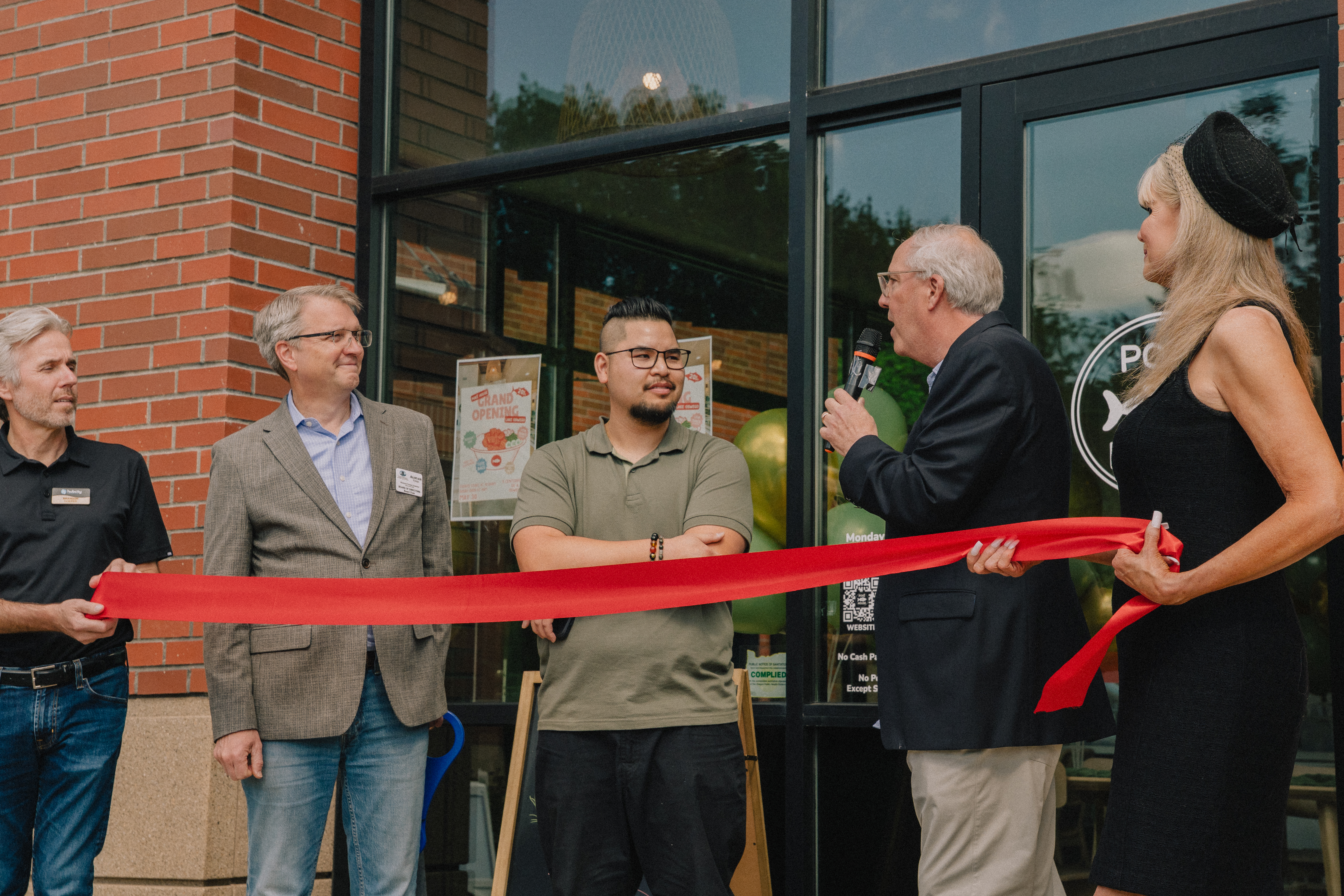 People standing at a ribbon-cutting ceremony outside a storefront, with one person holding a microphone and a red ribbon stretched in front.