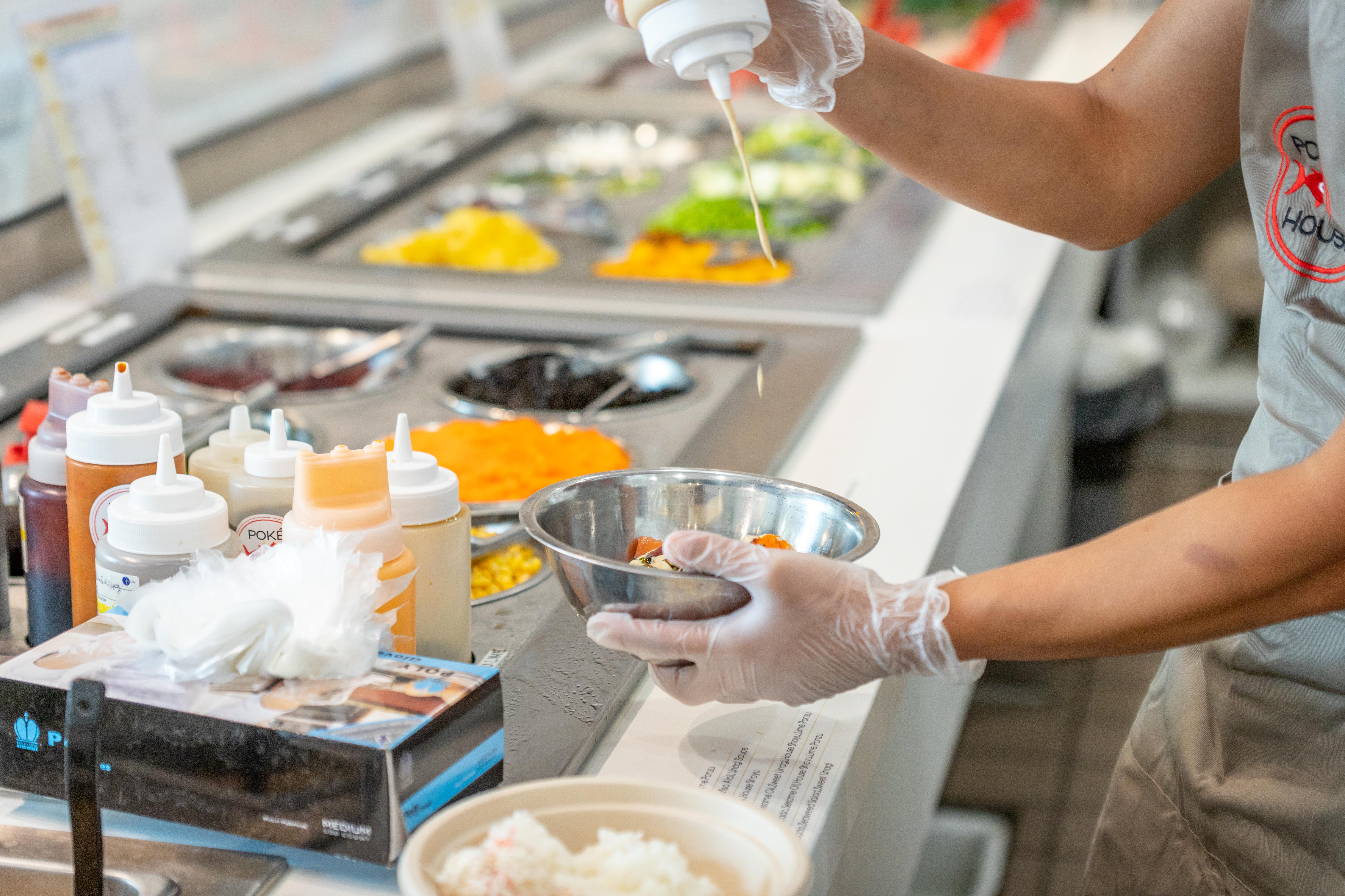 A person wearing gloves prepares food by pouring sauce into a metal bowl at a salad bar with various colorful ingredients.