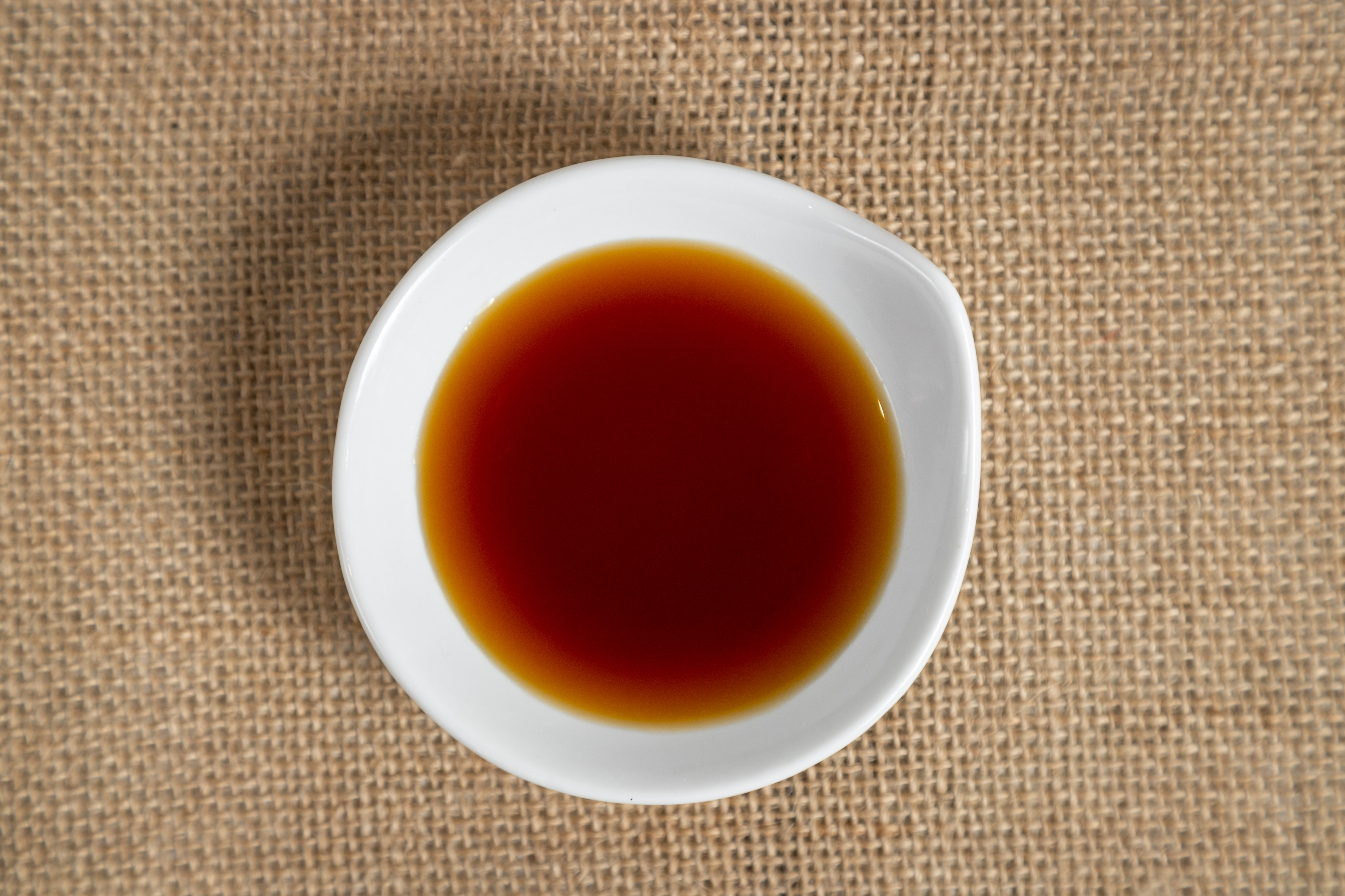 A small white bowl filled with dark brown liquid on a burlap surface.
