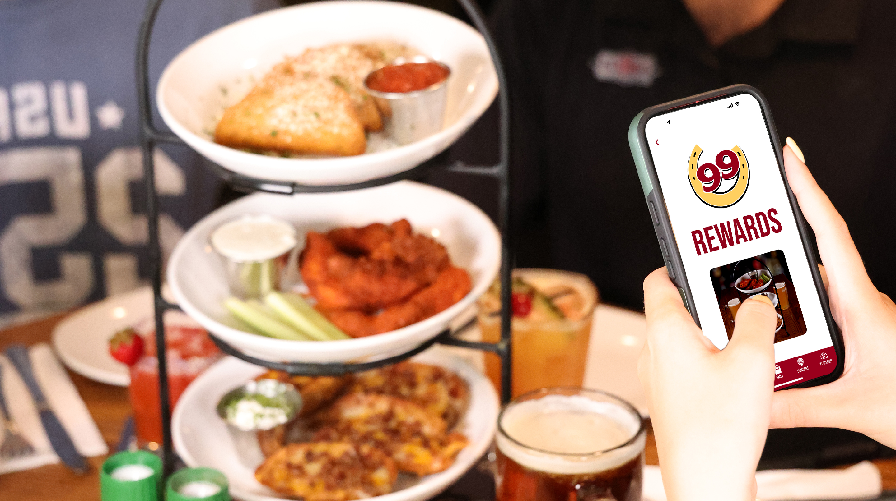 A set of hands holding a phone that is open on the 99 Restaurants app with a tower of food to the left behind it