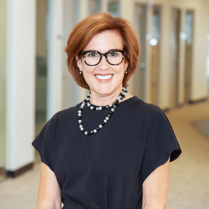 Smiling woman with short red hair and glasses wearing a black top and necklace, standing in a hallway.