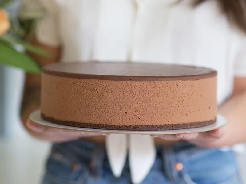 Woman in a white blouse holding a chocolate cake on a plate, smiling slightly, with blurred background.