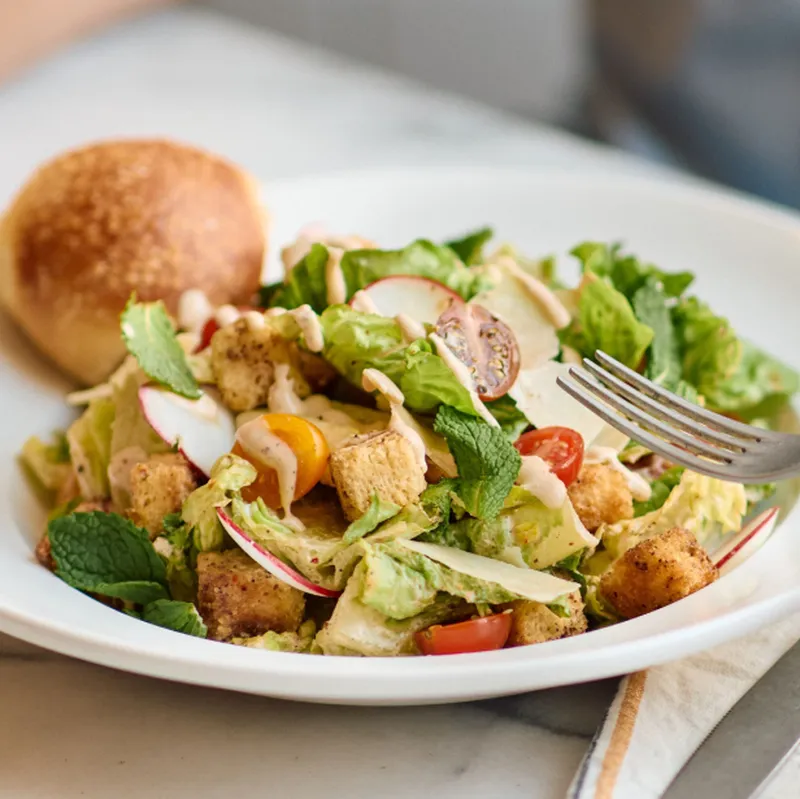 A fresh salad with lettuce, cherry tomatoes, radishes, croutons, and dressing, garnished with mint leaves. Served with a roll on a plate.