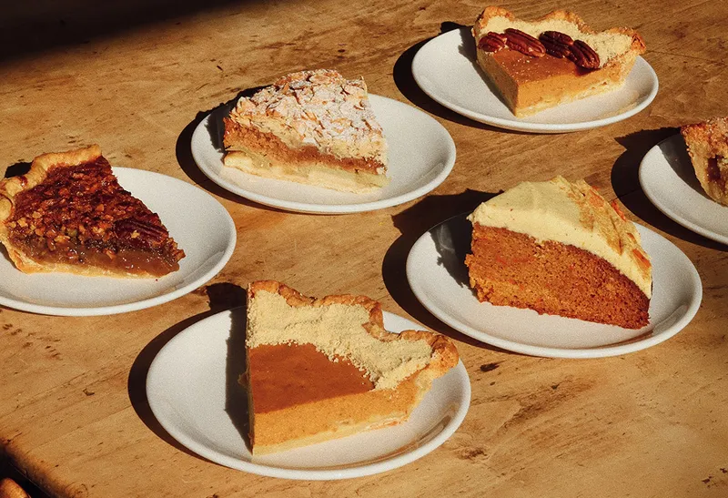 Various slices of pie arranged on white plates on a wooden table, including pecan, pumpkin, and carrot cake pie.
